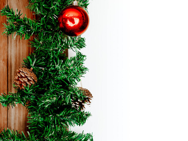 Christmas background with ornaments. Green fur decoration with red ball and pine cone on grunge brown wooden board and empty white background with copy space. - Photo, Image