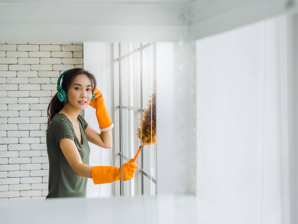 Krásná mladá Asiatka, žena v domácnosti v oranžových gumových rukavicích, poslouchá hudbu se zeleným sluchátkem a používá pírko čistící skleněné okno v bílém pokoji s kopírovacím prostorem. - Fotografie, Obrázek