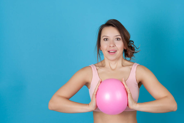 close-up portrait of a surprised girl in a pink sports top clutching a Pilates ball at her chest, isolated on a blue background - Photo, image