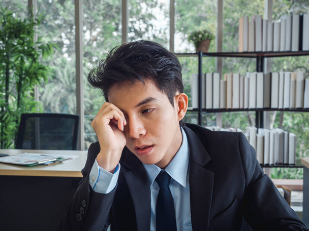 Jonge Aziatische zakenman in pak met problemen, moe, gestrest en verdrietig saai zitten met afwezig-minded op zijn bureau in het kantoor. Frustrated werknemer geestelijke gezondheidsproblemen en burn-out syndroom concept. - Foto, afbeelding