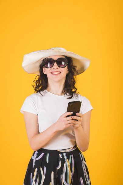 Beautiful young girl in sunglasses and a big white hat with a phone in her hands on a yellow background - Photo, Image