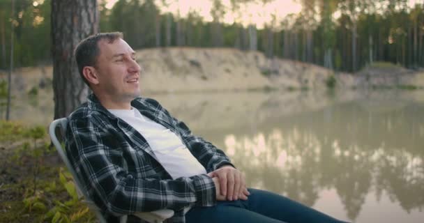 samotny człowiek relaksuje się na łonie natury, siedzi w fotelu nad brzegiem leśnego jeziora i cieszy się ciszą i spokojem - Materiał filmowy, wideo