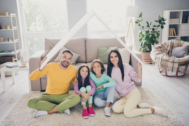 Photo of full family four members two little kids parents hold roof sit floor wear colorful sweater in living room indoors - Foto, Imagen