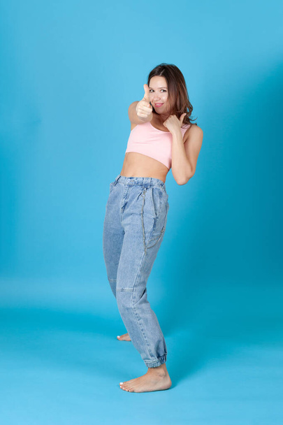 a full-length young woman in jeans pretends to shoot her fingers at us, isolated on a blue background - Photo, Image