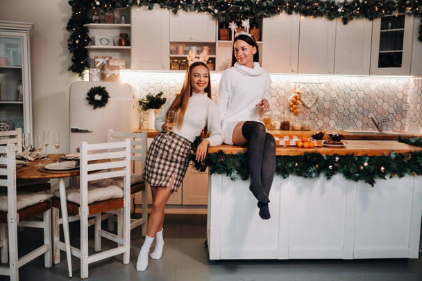 two girls in a cozy home environment in the kitchen with champagne in their hands for Christmas. Smiling girls drink champagne on a festive evening - Fotó, kép