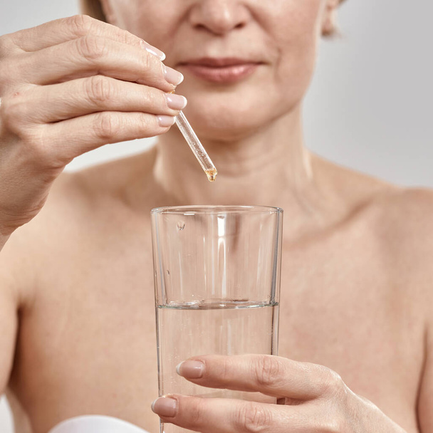 Close up of hand of middle aged woman holding dropper with medication and glass isolated over grey background - Valokuva, kuva