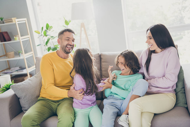 Photo of full big family four people sit sofa tickling having fun wear colorful pullover pants in living room indoors - Photo, image
