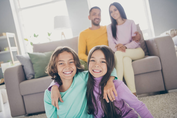 Photo of full family four members sit couch carpet two small kids twins embrace wear colorful pullover trousers in living room indoors - Fotografie, Obrázek