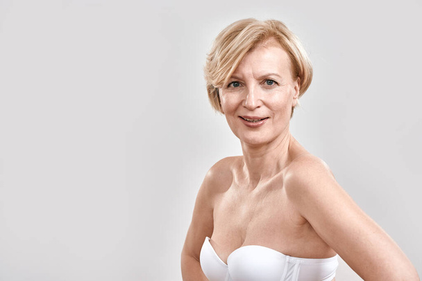 Portrait of beautiful middle aged woman in white bra looking cheerful at camera, posing isolated against grey background. Beauty concept - Фото, зображення