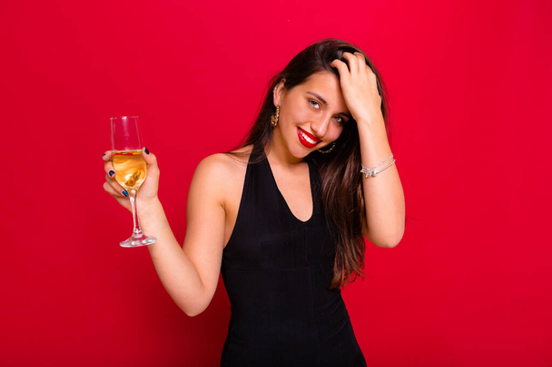 Mulher muito feliz endireita o cabelo com uma taça de champanhe usando roupas pretas sobre fundo vermelho. Foto de alta qualidade - Foto, Imagem