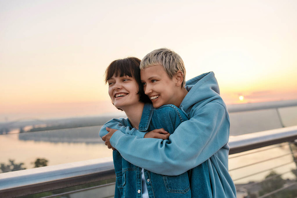 Young loving lesbian couple smiling, looking happy while enjoying romantic moments together, standing on the bridge at sunrise - 写真・画像