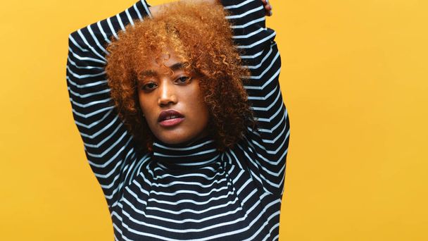 Young african american black woman with curly afro hair posing in front of yellow background with hands behind her head - 写真・画像