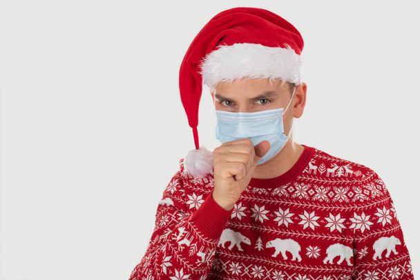 Portrait of young handsome caucasian man wearing Santa's hat, dressed with christmas outfit is posing on isolated with surgical mask - Fotó, kép