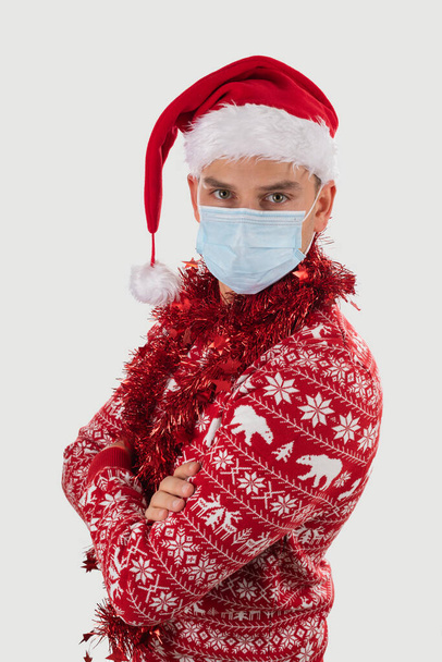 Portrait of young handsome caucasian man wearing Santa's hat, dressed with christmas outfit is posing on isolated with surgical mask - 写真・画像