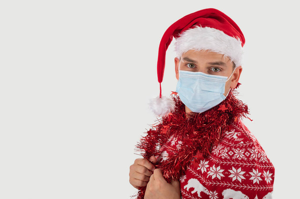 Porträt eines jungen gutaussehenden kaukasischen Mannes mit Weihnachtsmütze, gekleidet mit Weihnachtskostüm posiert isoliert mit Chirurgenmaske - Foto, Bild