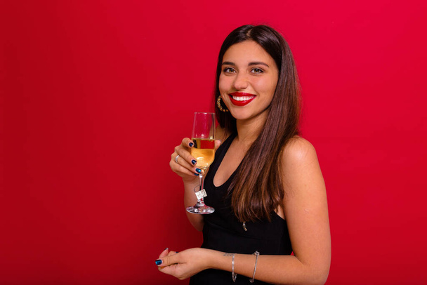 Happy pretty lady dressed black shirt with red lips is posing at camera with a glass of champagne over red background . High quality photo - Foto, Imagem