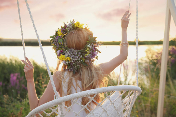 Young woman wearing wreath made of beautiful flowers on swing chair outdoors, back view - Fotografie, Obrázek