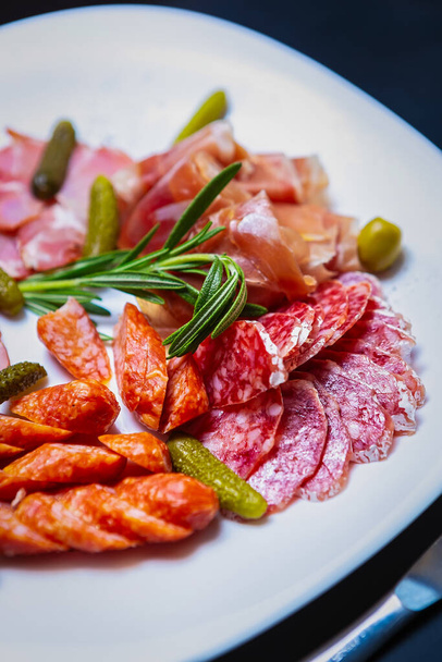 Delicious set of meat snacks in restaurant.Sliced Spanish jamon,smoked ham,pepperoni sausages served on white plate with pickled cucumbers,green olives.Gourmet wine appetizer foods in vertical photo - Photo, image
