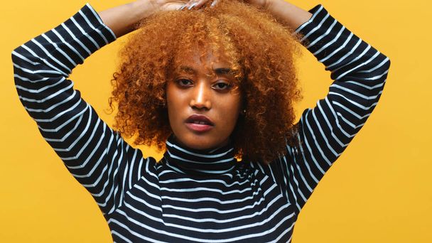 Joven afroamericana negra con el pelo rizado afro posando delante de fondo amarillo con las manos detrás de la cabeza - Foto, imagen