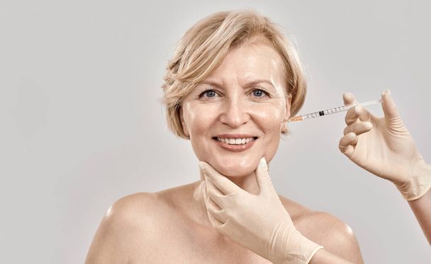 Portrait of beautiful naked middle aged woman smiling at camera while she gets injection in her face, posing isolated against grey background - Foto, immagini