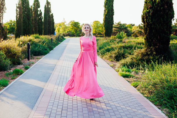 Kaunis blondi nainen vaaleanpunainen mekko kävelee puistossa auringonlaskun aikaan - Valokuva, kuva