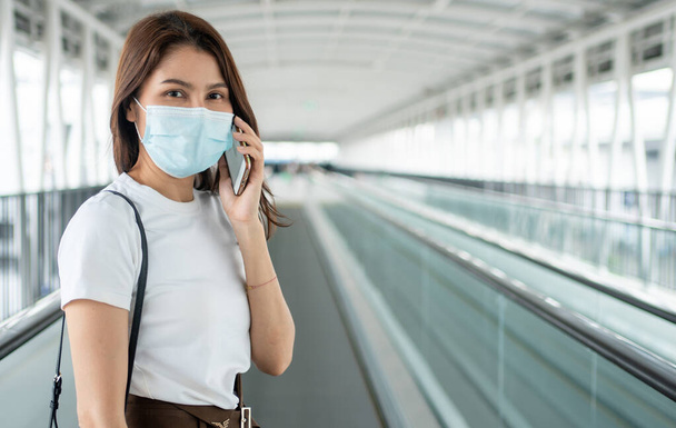 Портрет молодой женщины в медицинской маске для антикоронавирусной защиты от пандемических инфекционных заболеваний COVID-19 и использование смартфона в общественных местах. Концепция вирусной пандемии и загрязнения - Фото, изображение