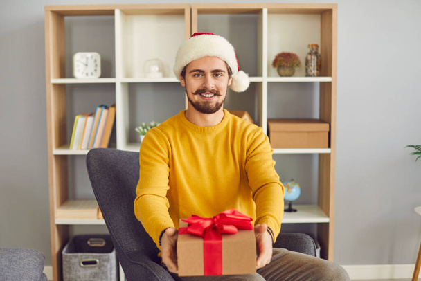 Young smiling man in festive Satna hat sitting showing holiday present box during videocall - Foto, imagen