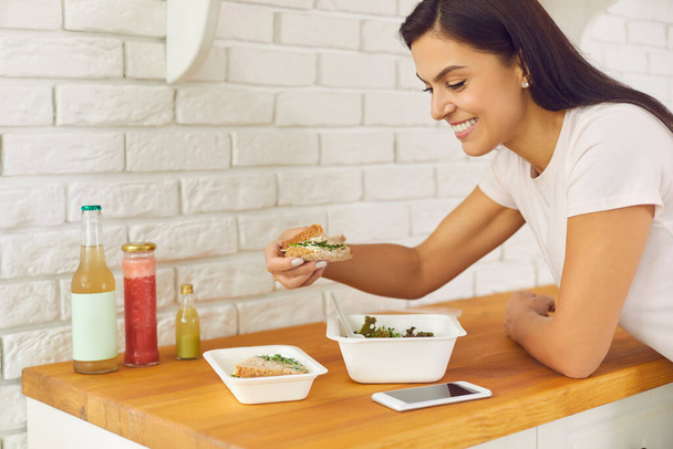 Happy young woman enjoying healthy takeaway vegetable salad and sandwich at home - Photo, image