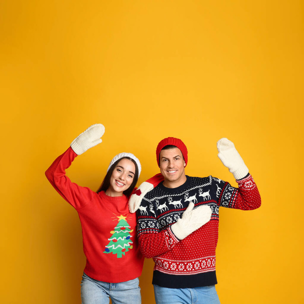 Couple in Christmas sweaters, knitted mittens and hats on yellow background, space for text - Fotoğraf, Görsel