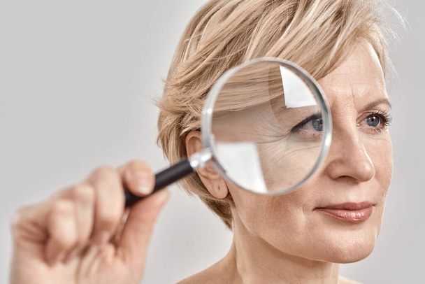 Close up portrait of attractive middle aged woman looking aside, holding a magnifying glass and showing her wrinkles, posing isolated over grey background - Foto, Imagem
