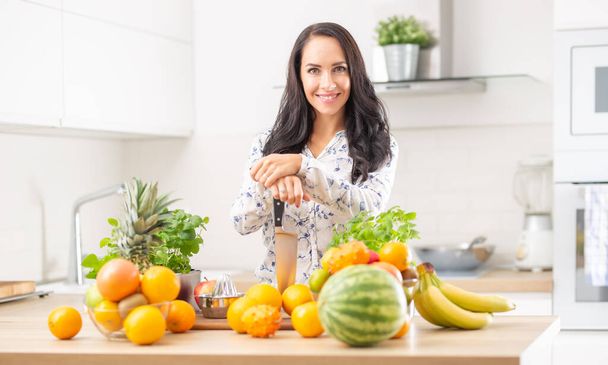 Smiling young woman holds a knife in the kitchen surrounded by fresh fruit and herbs. - 写真・画像