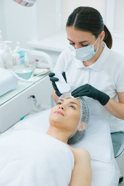 Mirthful woman smiling while a concentrated cosmetologist performing an ultrasonic cleansing procedure on her forehead - Zdjęcie, obraz