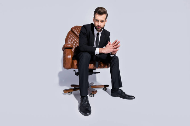 Full size low below angle view photo of handsome business guy sit office chair hold fingers together serious look wear black blazer pants tie shirt shoes suit isolated grey background - Фото, изображение
