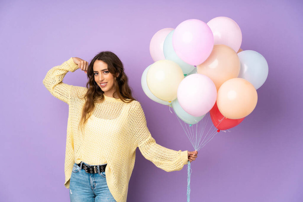 Mladá žena chycení mnoho balónů nad izolované na fialovém pozadí dělat silné gesto - Fotografie, Obrázek
