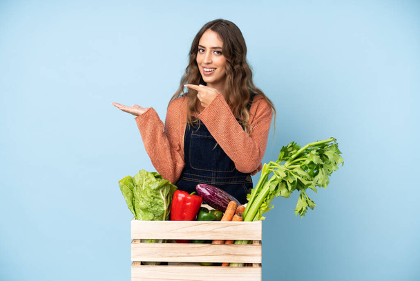 Frissen szedett zöldségekkel rendelkező gazda egy dobozban, aki a tenyerén képzeletbeli másolóteret tart, hogy reklámot helyezzen el - Fotó, kép