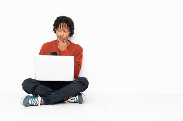 Młody Afroamerykanin siedzi na podłodze i pracuje z laptopem myśląc i wysyłając wiadomość - Zdjęcie, obraz