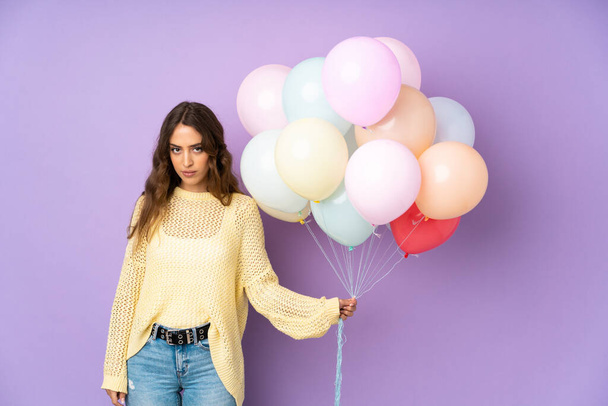 Young woman catching many balloons over isolated on purple background keeping arms crossed - 写真・画像