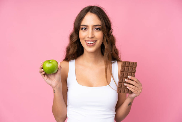 Young woman over isolated pink background taking a chocolate tablet in one hand and an apple in the other - Foto, Imagem