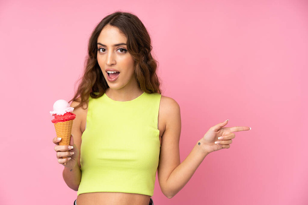 Jovem segurando um sorvete de corneto isolado em um fundo rosa surpreso e apontando o dedo para o lado - Foto, Imagem