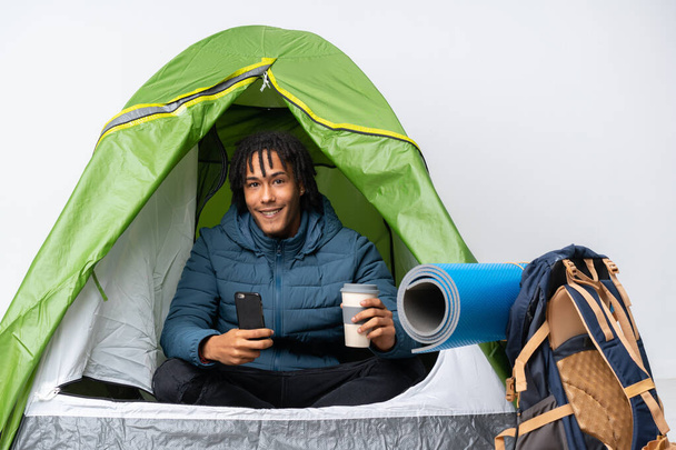 Young african american man inside a camping green tent holding coffee to take away and a mobile - Фото, зображення