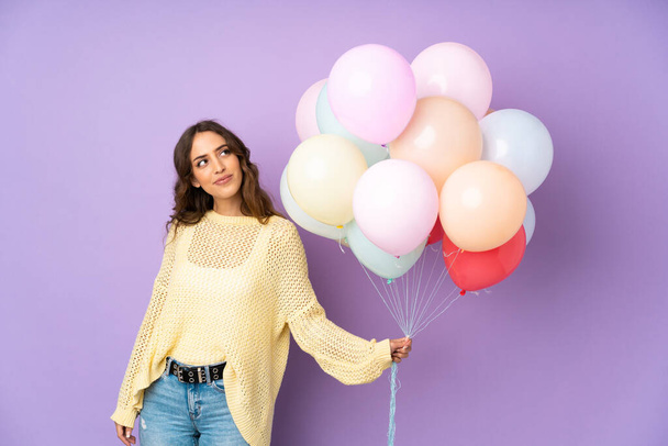Νεαρή γυναίκα αλίευση πολλά μπαλόνια πάνω απομονωμένο σε μωβ φόντο γέλιο και κοιτάζοντας ψηλά - Φωτογραφία, εικόνα