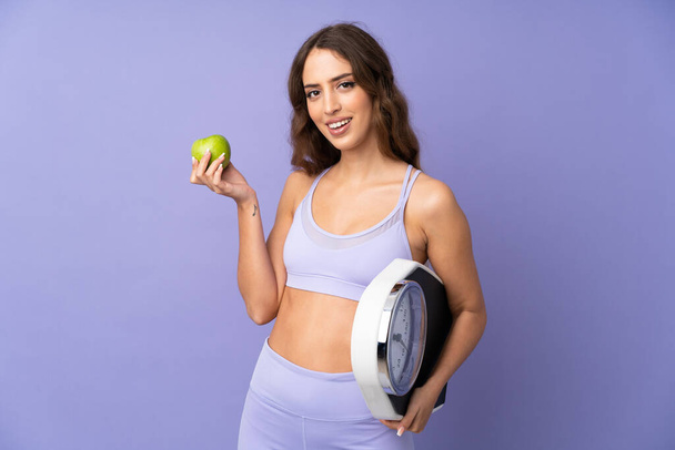 Young sport woman over isolated purple background with weighing machine and with an apple - Фото, зображення
