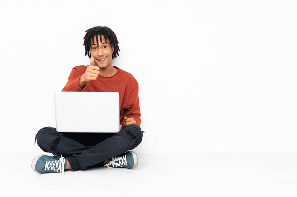 Fiatal afro-amerikai férfi ül a földön, és hüvelykujjal feltartott laptoppal dolgozik, mert valami jó dolog történt. - Fotó, kép