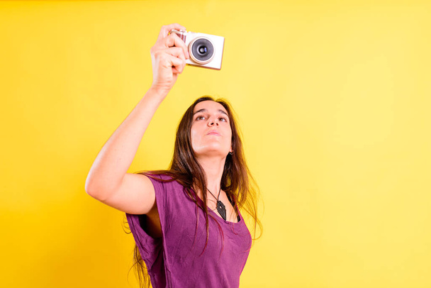 Ładna młoda dziewczyna robi zdjęcia za pomocą złego aparatu, odizolowany na żółtym tle studio. - Zdjęcie, obraz