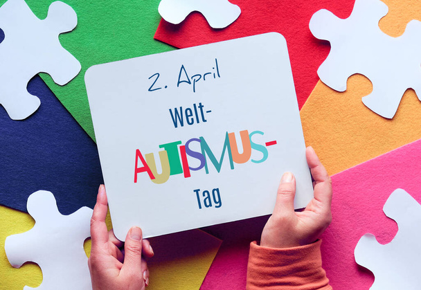 Welt-Autismus-tag design. Hands holding page with text in German that means April 2 Autism World Awareness day. Top view on jigsaw puzzle element on layered colorful felt background. - Photo, image