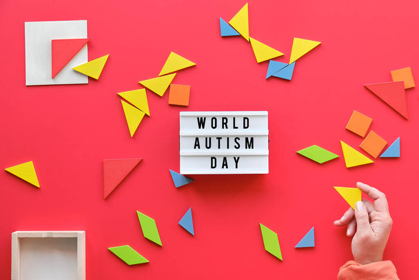 Creative design for Autism World Day on April 2, text on lightbox. Tangram elements scattered on red background. Hand holding triangle. - Zdjęcie, obraz