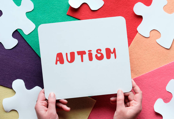 Concept paper design, World Autism Awareness Day. Jigsaw puzzle elements on felt pieces, top view. Hands hold cardboard placard with text Autism.. - Fotografie, Obrázek