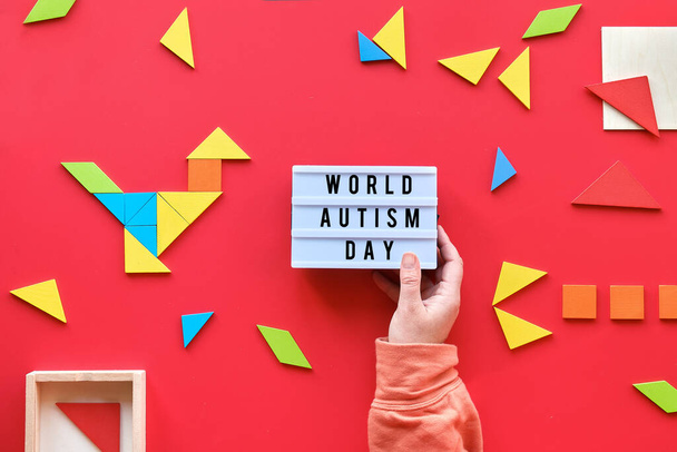Creative design for Autism World Day on April 2, text on lightbox. Tangram puzzle elements scattered on red background. Hand hold light box. - Foto, imagen