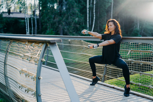 Motivated brunette active young woman does squat exercises with dumbbells, trains biceps, dressed in black active wear, poses at bridge outdoor during sunrise, has morning workout. Sport concept - Photo, Image