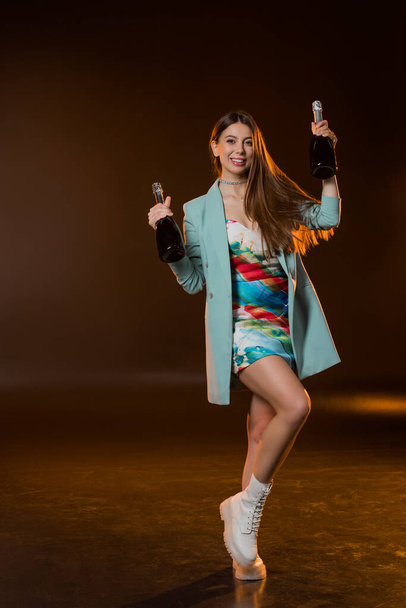 full length of cheerful brunette woman in jacket holding bottles with champagne on black  - Fotografie, Obrázek